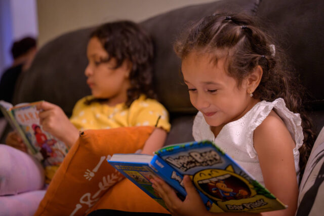 Two girls sit on a couch reading books.