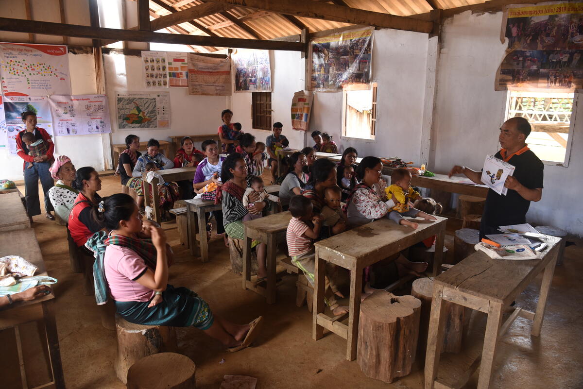 Teacher reading a book to attentive students in a classroom filled with wooden desks and educational posters.