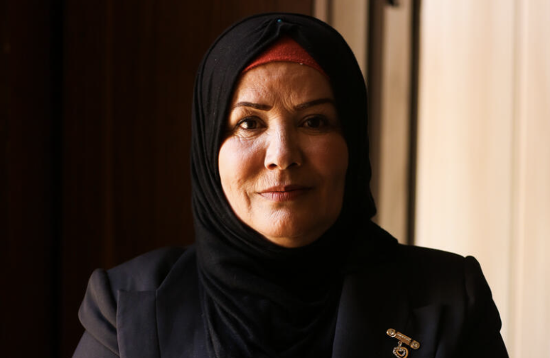 A confident woman smiles slightly while wearing a hijab and blazer.