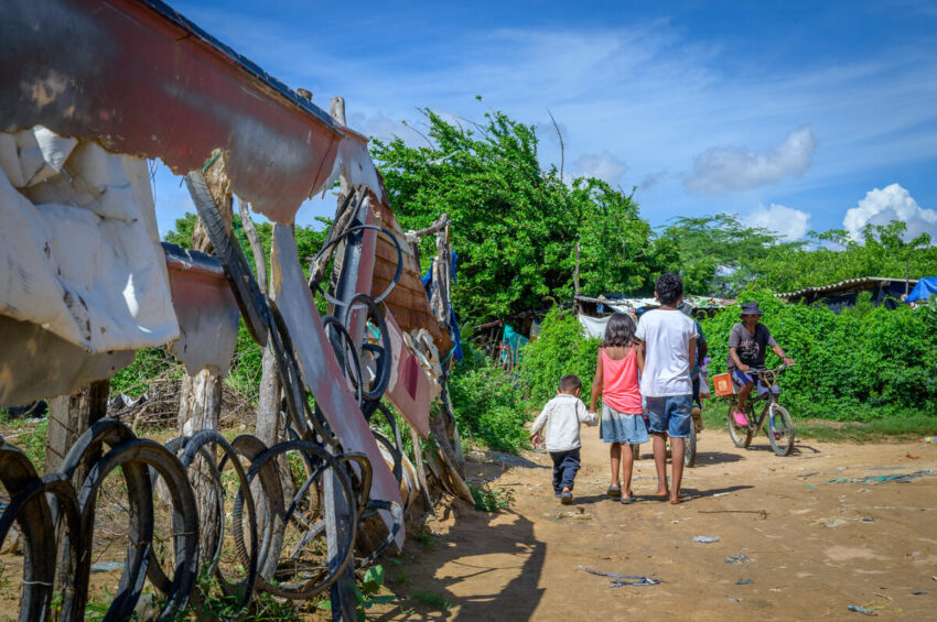 Children hold hands and walk away from the camera along a dirt road with bicycle tires and tattered siding hanging from posts on the left. A cyclist enters the scene.