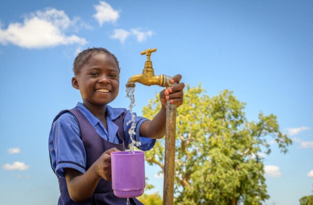 A smiling girl holding a large purple mug under a water source with water running from a faucet. In the background, a tree and a bright blue sky with white clouds.