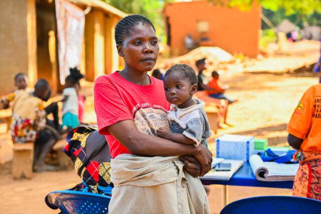 A woman in a red T-shirt holds a child on her hip, gazing away from the camera. Sunlight shines, with a table behind her.