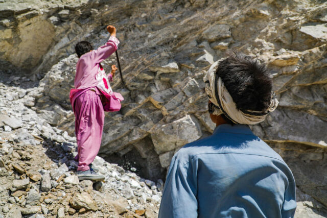 A young boy, wearing dark pink, digs with a shovel into a rocky hillside, while another boy, dressed in blue with a white scarf headband, watches him.