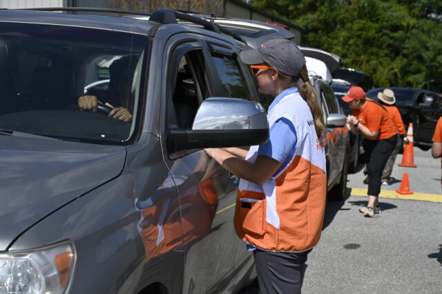 A woman wearing a World Vision vest and baseball cap speaks with the motorist. A line of cars and people in orange are in the background.