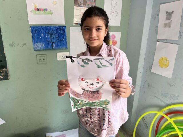 A girl wearing a pink blouse smiles at the camera, holding a handmade picture of a smiling white cat on grass with a tree branch above.