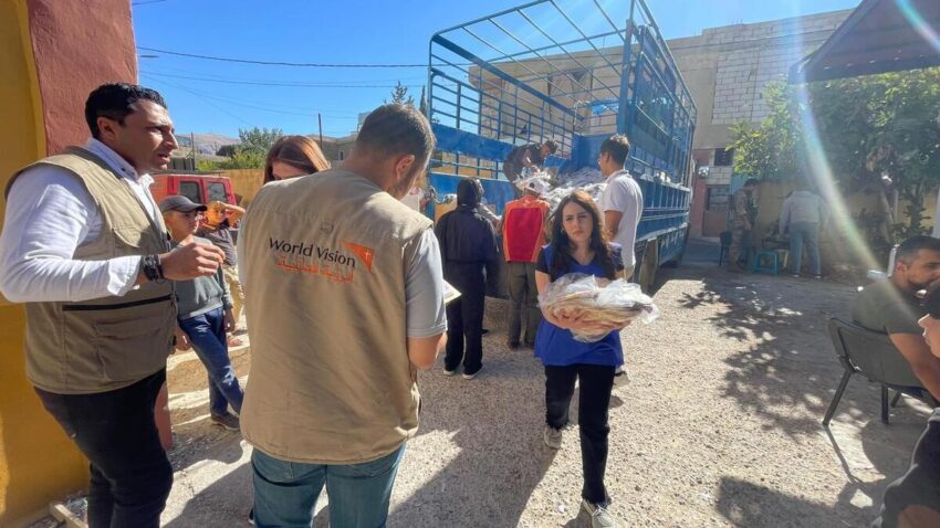 A woman in a blue vest carrying bread walks toward a man with a World Vision vest. A truck with blue siding and a brick are visible in the bright sun.