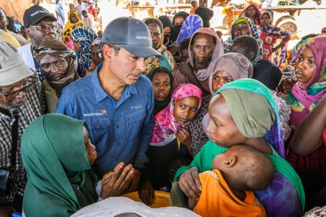 A man in a World Vision shirt and hat is surrounded by a crowd of people, many looking distressed. He speaks to a woman holding a young child.