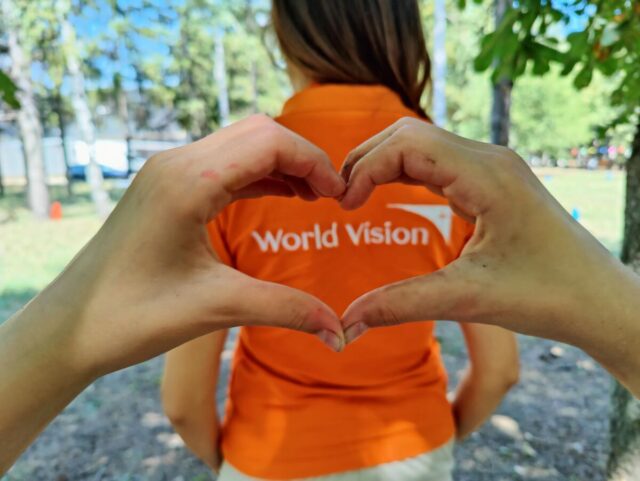 Two hands form a heart shape around the back of a woman in an orange World Vision T-shirt, with blurred trees and sunlight in the background.