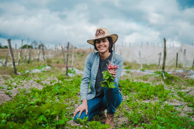 A woman kneels in a garden of greens while holding a bunch of turnips. She is smiling, and there are more gardens behind her.