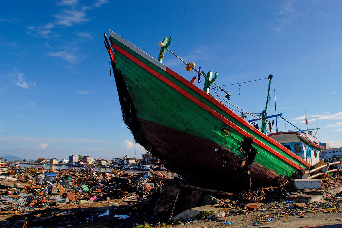 The large green hull of a ruined boat projects upward among the tsunami-devastated ruins of a coastal city, surrounded by debris under a blue sky.