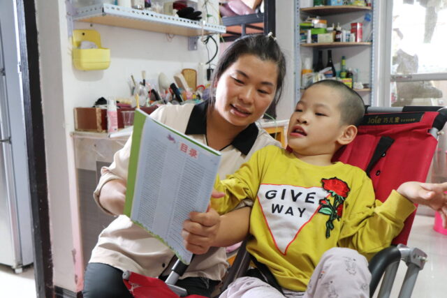 A woman kneels next to a young girl in a wheelchair and holds a book up in front of her, both looking at the page.