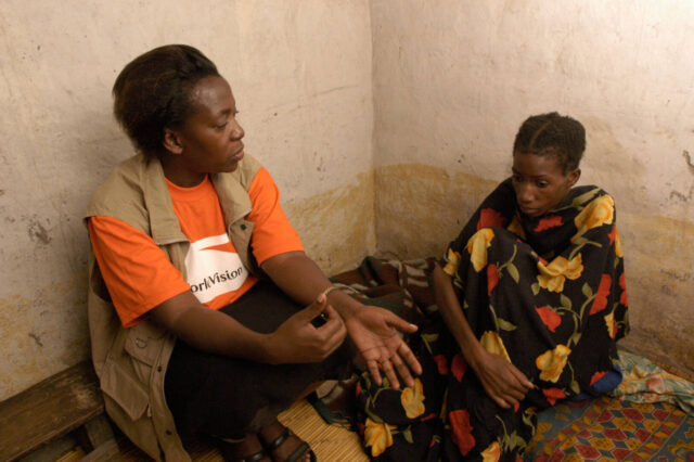 A woman in a World Vision shirt and vest sits on the floor next to a frail woman.