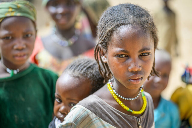 A Sudanese girl wearing colored beads looks wearily into the camera while carrying a young child on her back. Other children are behind them.