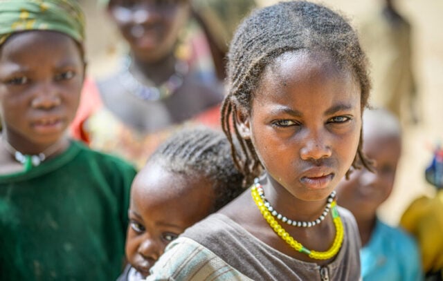 A Sudanese girl wearing colored beads looks wearily into the camera while carrying a young child on her back. Other children are behind them.