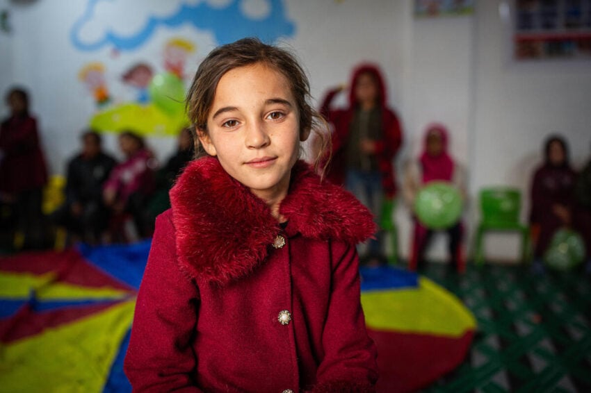 A girl with her hair pulled back wears a red coat with a faux fur collar and gazes directly at the camera. Blurred children sit and stand in the background.
