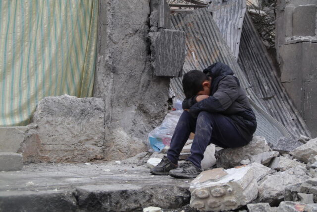 A boy sits on top of rubble with his head resting on his knees. Behind him is a collapsed building with aluminum sheets and a hanging blanket.