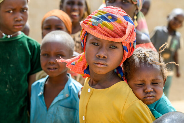 A girl with a vibrant patterned orange headscarf and yellow button-down shirt gazes at the camera while carrying a baby on their back. Children surround them.