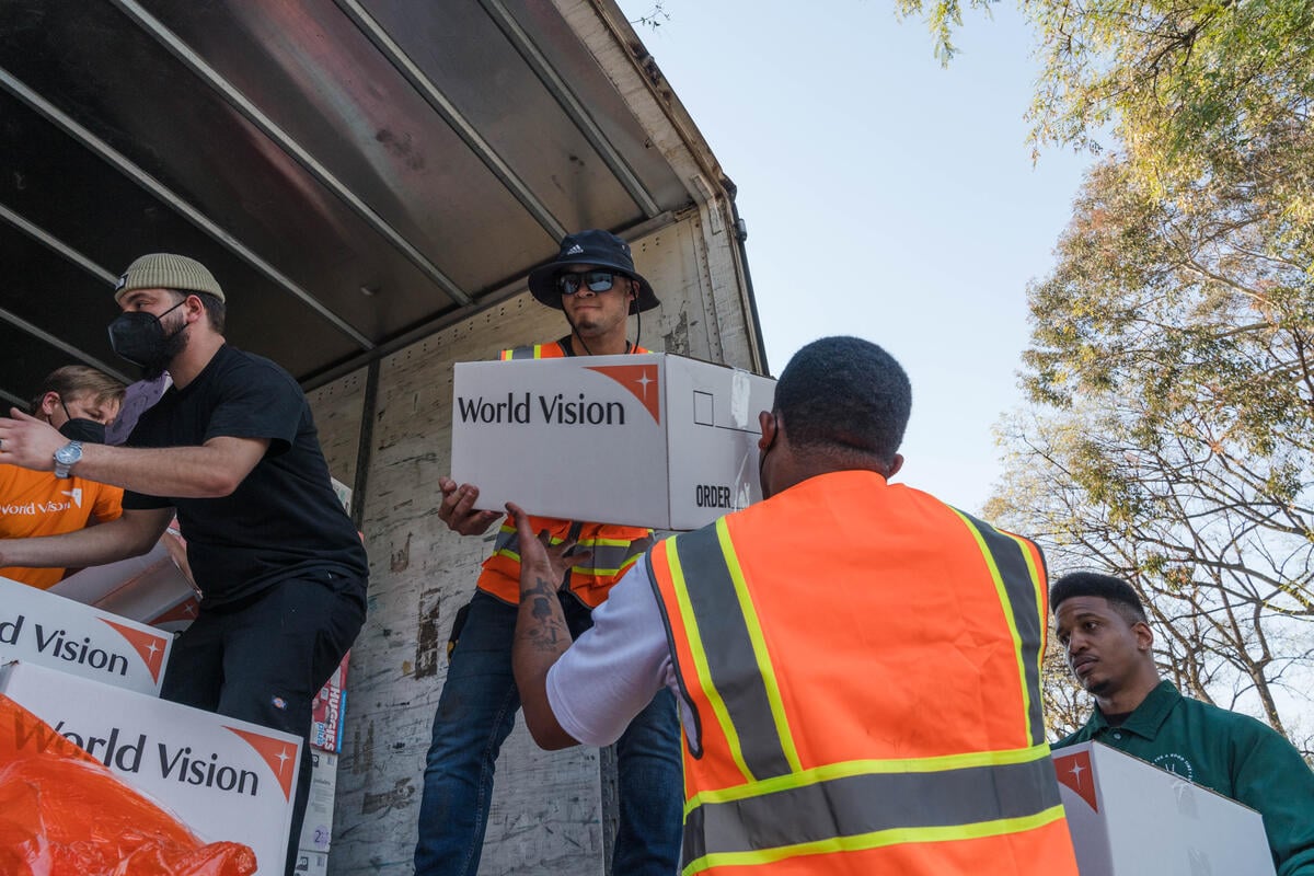 Men unload boxes of disaster relief supplies from a truck during an aid distribution event on a sunny day.