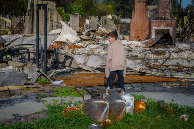 A boy wearing an oversized jacket walks through the ruins and debris of his family’s home following a devastating wildfire.