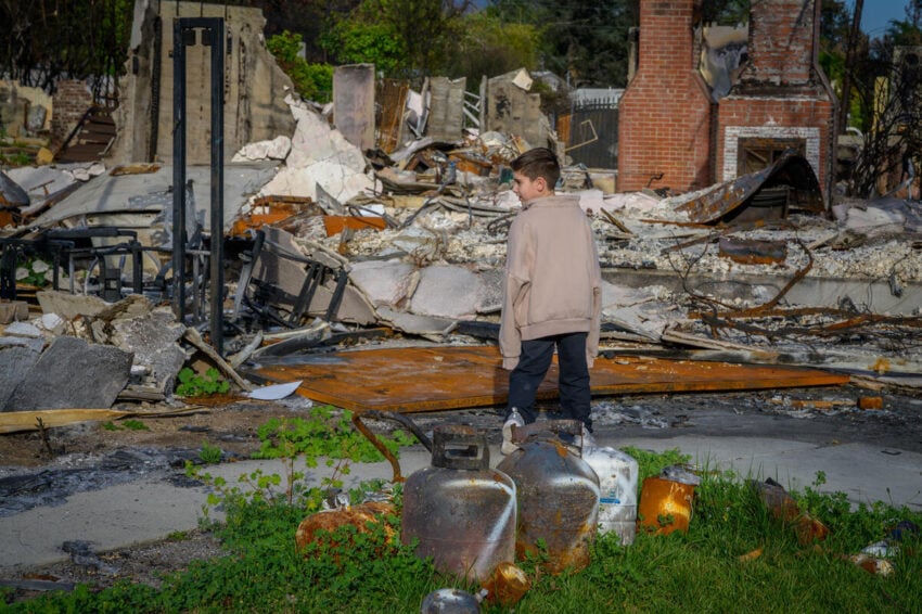 A boy wearing an oversized jacket walks through the ruins and debris of his family’s home following a devastating wildfire.