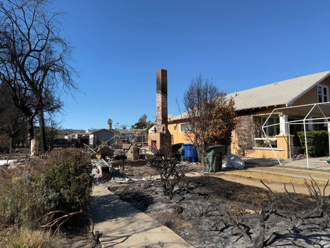 A brick fireplace and chimney are all that stands of a house burned down, while a yellow house next door has minimal damage.