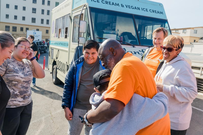 People look on as a man embraces a child. An ice cream truck is behind them.