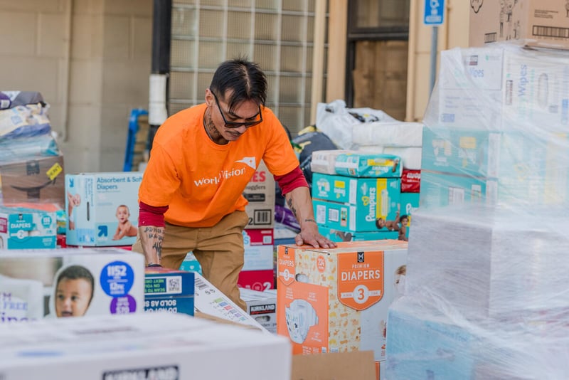 A man tilts a box up while holding another in place. He is surrounded by piles of packages of diapers as tall as he is.