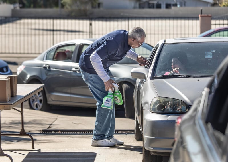 A tall older man bends down to greet people in a car while another car waits behind it. He is holding bottles of Lysol cleaner.