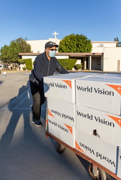 A man pushes a cart with more than 12 boxes that have the World Vision logo. A church with a cross on the roof is behind him.