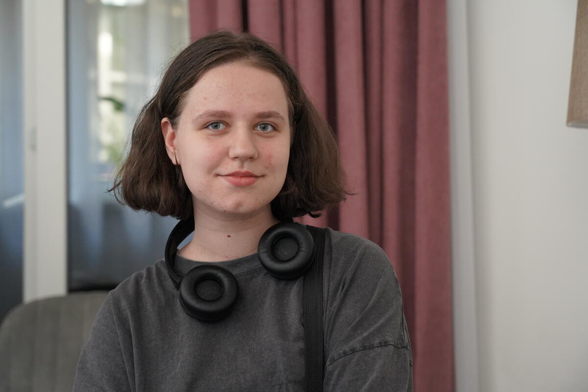 A young woman with short brown hair wearing black headphones around her neck, sitting in front of a pink curtain.