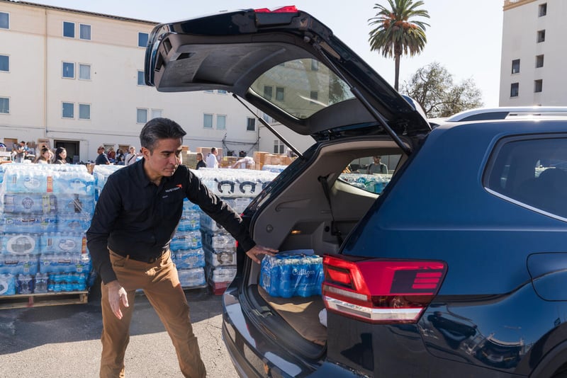 A man puts a case of bottled water in the back of a car. There are cases of water piled up behind him and people in the background.