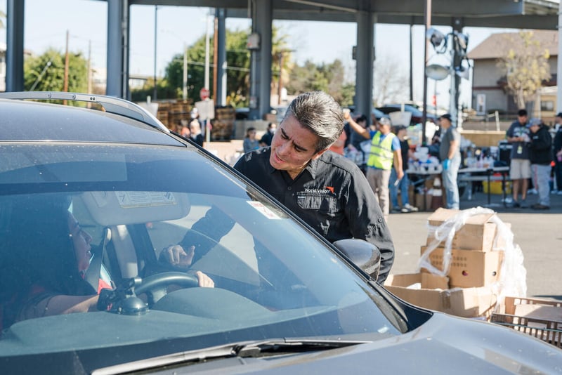 A man rests his arm on the partially open window of a car while talking to a woman driving it. There are boxes and people behind him.