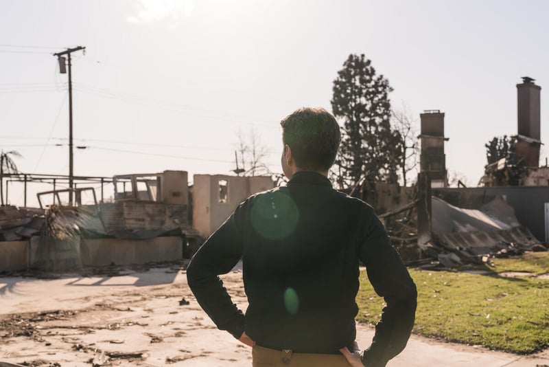 A man has his back to the camera and is looking out at burned buildings in front of him.
