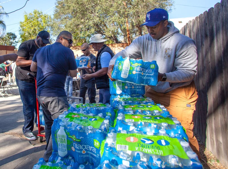 Men lift cases of water that will be distributed to people. There is a fence to the right and trees in the distance behind them.