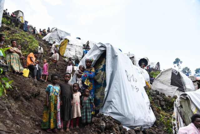 A hillside is covered with tents and makeshift shelters, with women and children scattered across the steep, terraced slopes, with people visible to the ridge line.