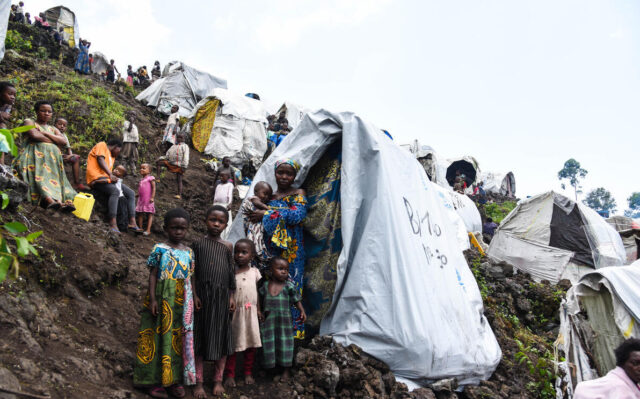 A hillside is covered with tents and makeshift shelters, with women and children scattered across the steep, terraced slopes, with people visible to the ridge line.