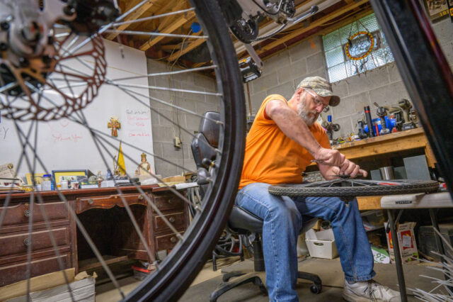 A man sits and works on a bicycle tire with a bicycle on a rack in front of him.