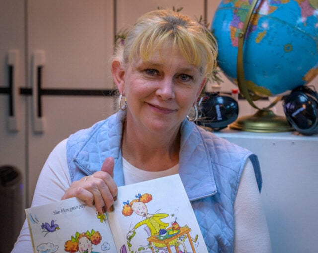 A woman sitting in front of a globe and cabinets holds a picture book in her hands.