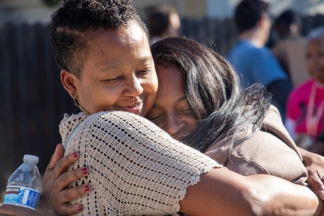 Two women, both with their eyes closed, hug each other. One holds a water bottle. There are people in the background.