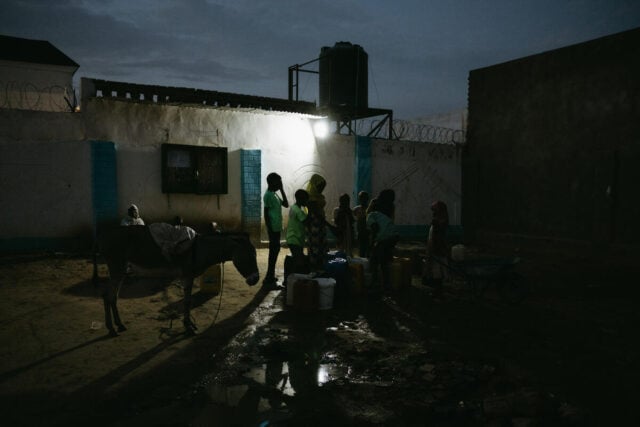 A light illuminates a dark scene where silhouetted children gather around jerry cans and a water tap.