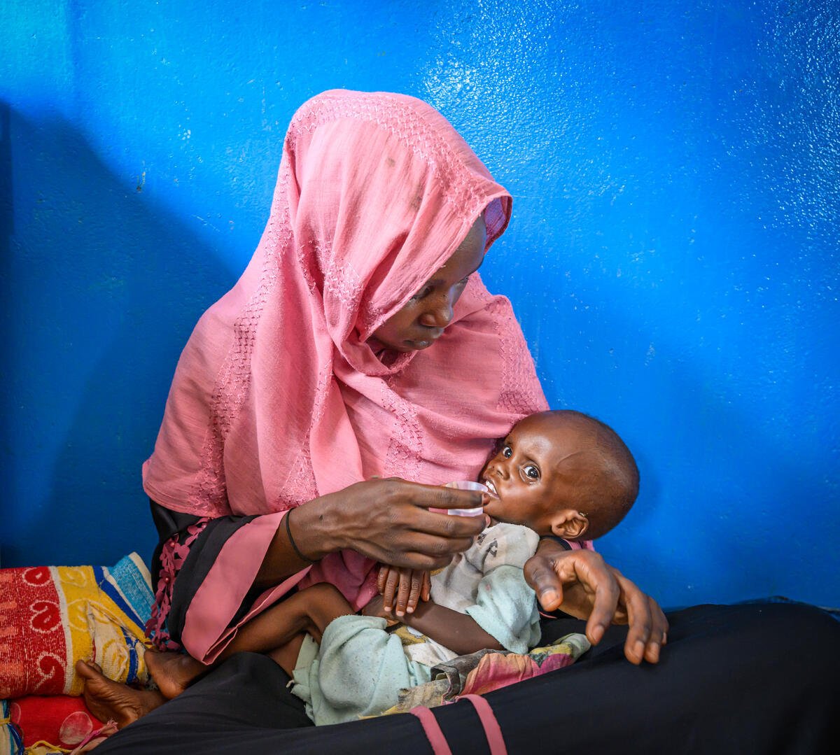A woman wearing a pink headscarf offers a cup to her thin, malnourished child, who looks toward the camera.