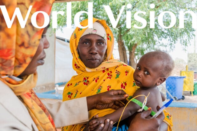A woman in a yellow patterned wrap holds a small child while a health worker uses a middle-upper arm circumference band to measure the child’s arm.