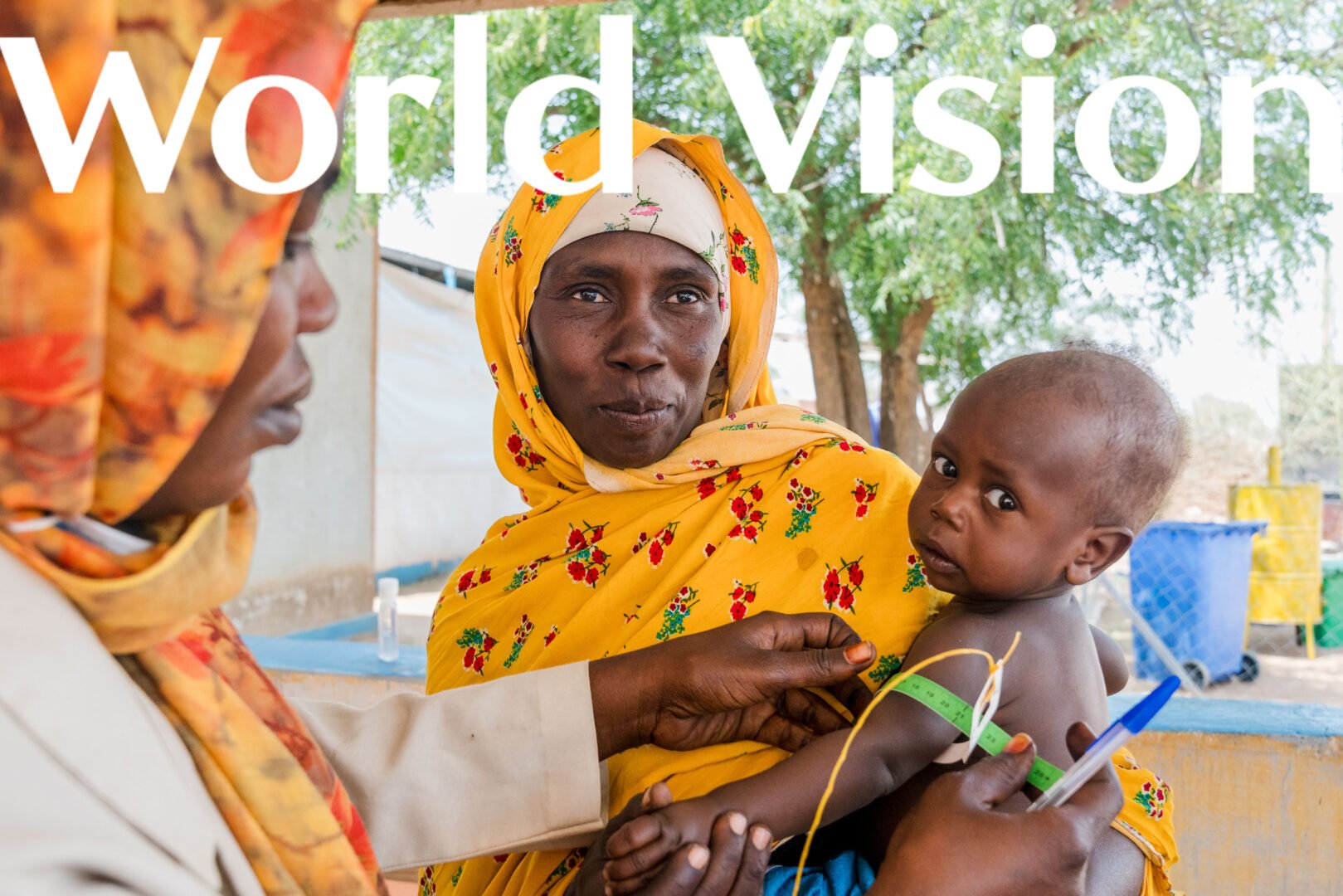 A woman in a yellow patterned wrap holds a small child while a health worker uses a middle-upper arm circumference band to measure the child’s arm.