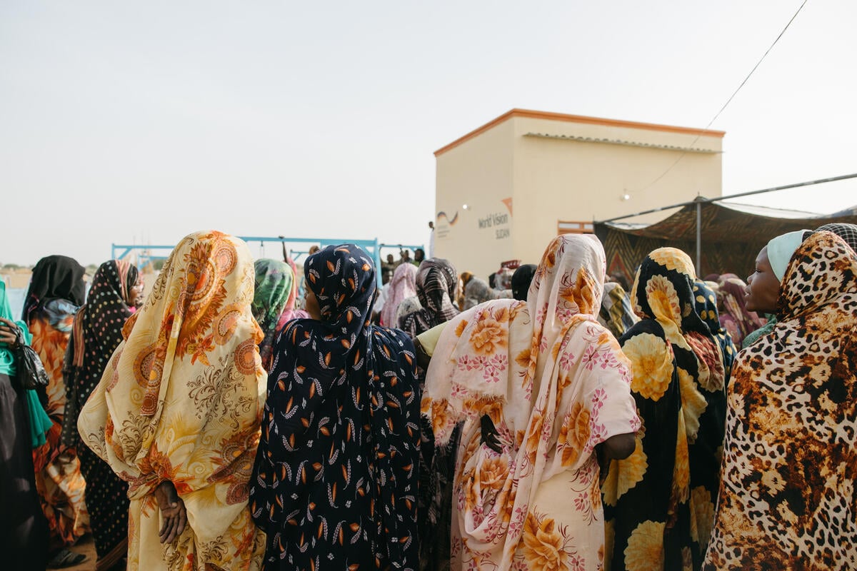 Women wearing colorful headscarves look toward a play area beside a World Vision building, where displaced children gather in a safe environment.