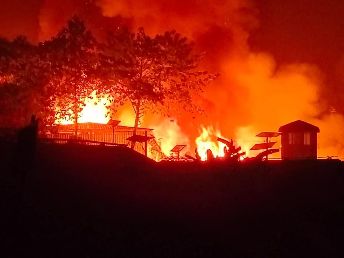 Flames and smoke rise into the night sky, silhouetting structures in a refugee camp. 