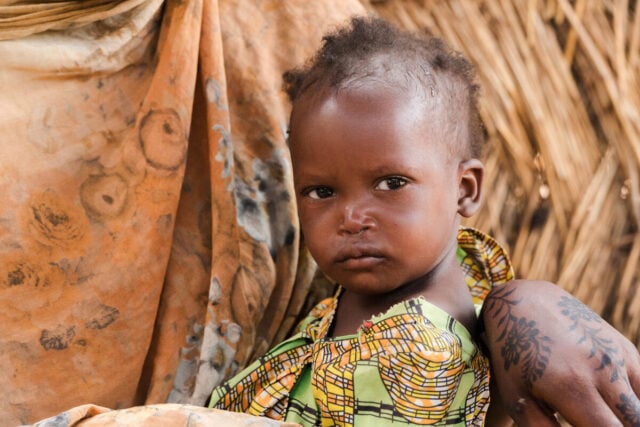 A young child sits on a woman’s lap, gazing into the camera. Behind them stands a makeshift shelter constructed from sticks.