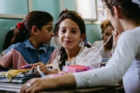 A young girl sits behind a classroom desk with books and colorful items while other girls surround her, glancing at the camera.