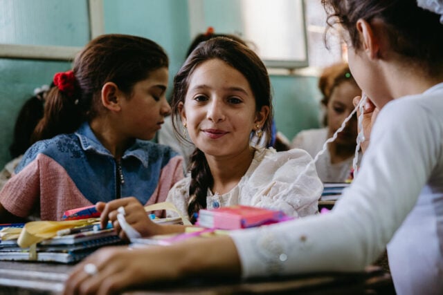 A young girl sits behind a classroom desk with books and colorful items while other girls surround her, glancing at the camera.