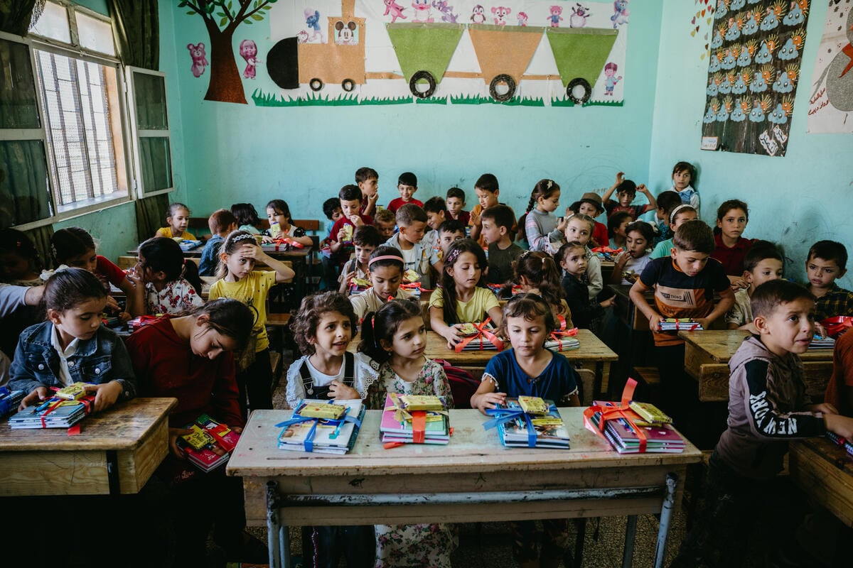 Children sit at desks in rows in a crowded classroom with colorful pictures on the wall. 