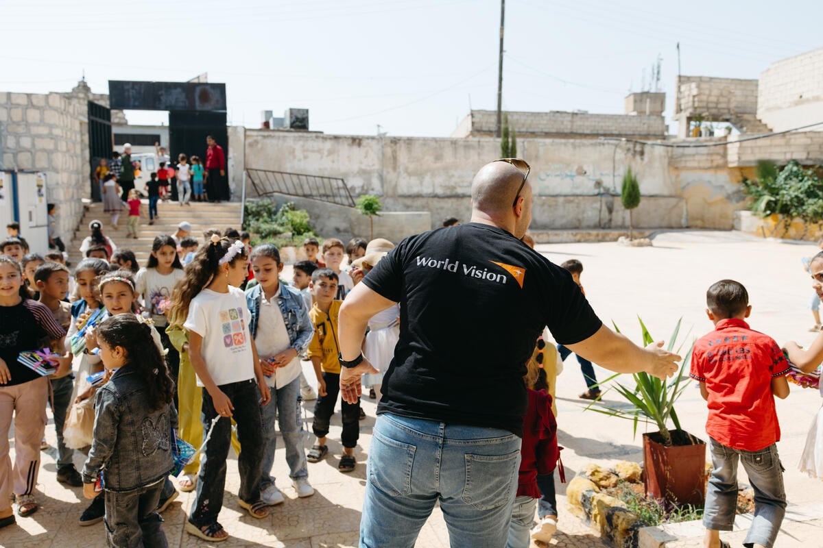 An aid worker wearing a World Vision T-shirt stands with his back turned from the camera. Children gather in a courtyard at a school in Syria.